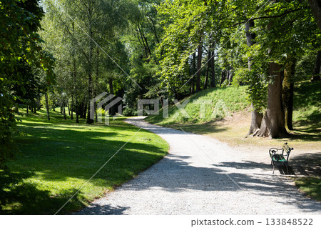 Tranquil park with a winding gravel path surrounded by lush green trees and a solitary bench. Peaceful nature scene bathed in warm sunlight 133848522