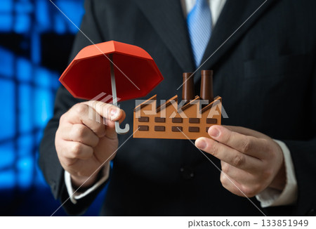 A businessman insures his factory with an umbrella. Insurance in industry. Protection of assets and risk management. Industrial safety, business continuity and corporate security policies. 133851949