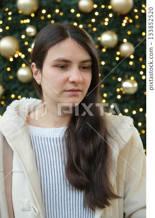 Thoughtful Young Woman in Cozy Coat Against Sparkling Christmas Tree Background. Thoughtful Young Woman in Cozy Coat Against Sparkling Christmas Tree Background. 133852520