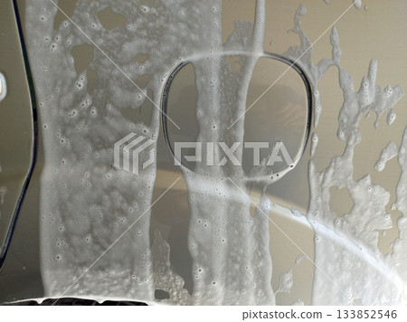 A close-up view of a cars fuel door covered in thick, white soap suds. The shiny surface and bubbly texture suggest the car is being washed 133852546