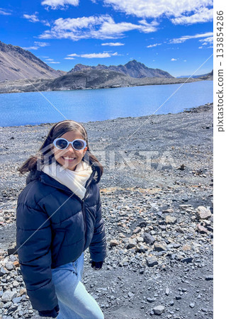 A woman wearing sunglasses and a black jacket stands in front of a body of water A woman wearing sunglasses and a black jacket stands in front of a body of water 133854286