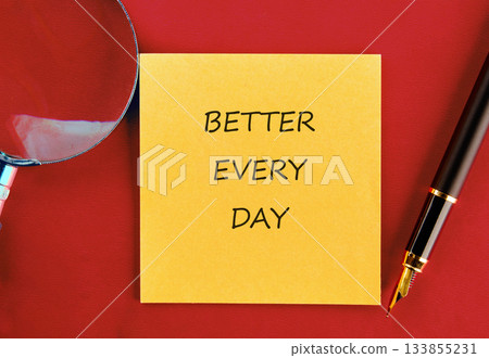 A bright yellow sticky note sits on a rich red surface, featuring the words Better Every Day. Nearby, a fountain pen and magnifying glass invite reflection and focus on personal growth 133855231
