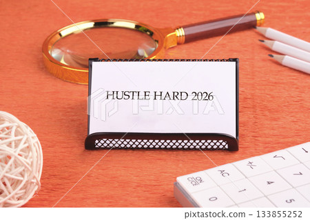On a warm wooden table, a motivational card reads Hustle Hard 2026, surrounded by a magnifying glass, pencils, and a unique decorative ball, symbolizing focus and creativity On a warm wooden table, a motivational card reads Hustle Hard 2026, surrounded by a magnifying glass, pencils, and a unique decorative ball, symbolizing focus and creativity 133855252