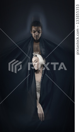Stylized portrait of two dancers wrapped in dark fabric on deep shadow background. 133855353