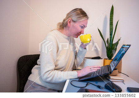 Woman drinking from mug while working on laptop at home office desk 133855602