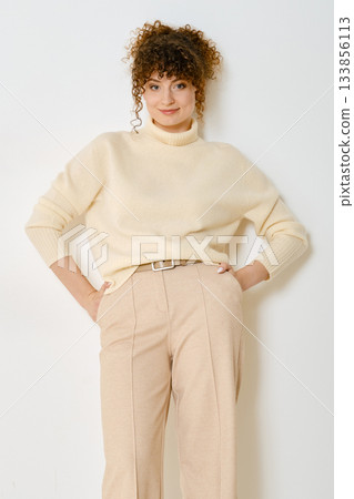 Stylish woman in cream sweater and beige pants poses confidently against a white background Stylish woman in cream sweater and beige pants poses confidently against a white background 133856113