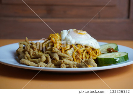 Delicious Mie Goreng Kari or Indonesian Curry fried noodle with crispy fried chicken offal, sunny-side-up egg, and cucumber slices on an orange background. Delicious Mie Goreng Kari or Indonesian Curry fried noodle with crispy fried chicken offal, sunny-side-up egg, and cucumber slices on an orange background. 133856522