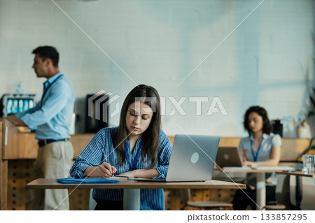 Female Student Taking Notes from Online Lecture in Sunny University Cafeteria During Lunch 133857295
