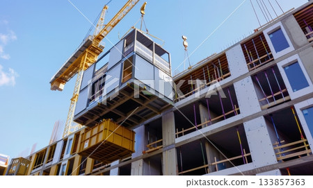 A construction site with a crane lifting a modular building unit. The structure is partially completed with exposed frames and windows. A construction site with a crane lifting a modular building unit. The structure is partially completed with exposed frames and windows. 133857363