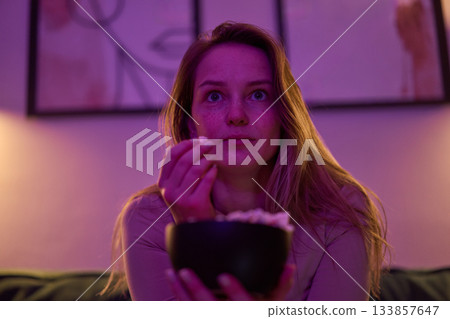 Female watching screen with popcorn bowl on sofa indoors Female watching screen with popcorn bowl on sofa indoors 133857647
