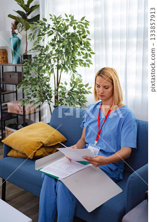 Female Doctor Reading Medical Records Sitting On Sofa At Home 133857913