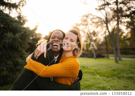 Two happy diverse women hugging in park during sunset Two happy diverse women hugging in park during sunset 133858012