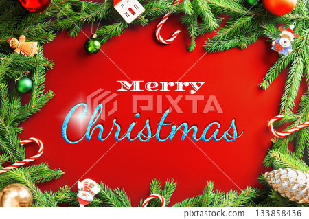 A vibrant red Christmas background framed by green fir branches and festive holiday ornaments with "Merry Christmas" text. High quality photo 133858436