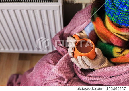 A close-up of a girl holding a cup of tea. She sits near a radiator. She's wearing a scarf, gloves, and a sweater. She's wrapped in a purple blanket. She's trying to keep warm in a poorly heated room, 133860337