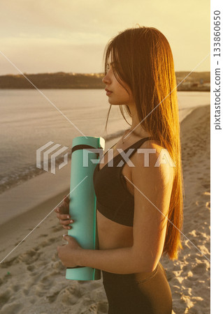 Woman holding yoga mat by the sea at sunset, fitness lifestyle, yoga training, outdoor wellness, healthy routine, seaside workout, calm evening yoga vibes. Woman holding yoga mat by the sea at sunset, fitness lifestyle, yoga training, outdoor wellness, healthy routine, seaside workout, calm evening yoga vibes. 133860650