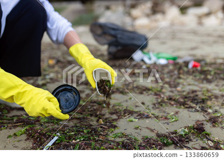Hands in yellow gloves collecting plastic cup waste during beach cleanup, highlighting pollution, environmental protection and ocean sustainability. 133860659