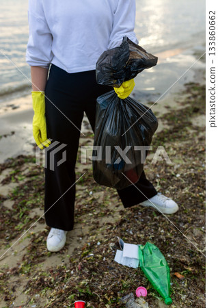 Volunteer collecting beach litter into a black trash bag during coastal cleanup, highlighting pollution, waste problem, environmental protection and eco awareness. 133860662