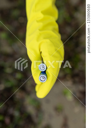 Close-up of a volunteer wearing yellow gloves holding discarded batteries collected on a polluted beach. Environmental cleanup, toxic waste, coastline protection and eco awareness. 133860680
