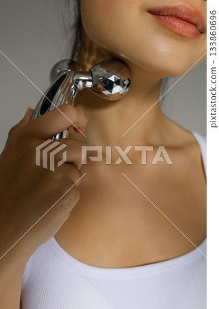 Close-up of a young woman massaging her neck with a metal 3D roller, demonstrating a tightening and relaxing skincare routine at home. 133860696