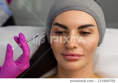 Close up of hands of young cosmetologist injecting botox in female face. She is standing and smiling. The woman is closed her eyes with relaxation 133860700