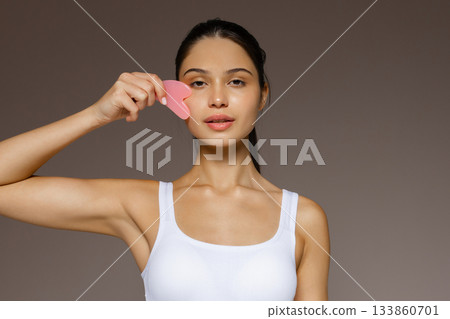 Young woman gently massaging her face with a pink gua sha stone, demonstrating a skincare routine against a neutral background. 133860701