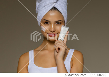 Young woman with a towel on her head applying skincare cream and holding a cosmetic tube, demonstrating a daily beauty routine. Young woman with a towel on her head applying skincare cream and holding a cosmetic tube, demonstrating a daily beauty routine. 133860702