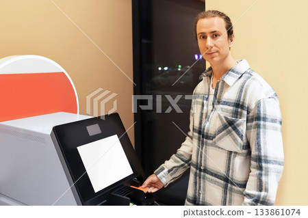 A young man in a plaid shirt uses a self-service terminal, inserting a card, possibly at an airport or station. High quality photo 133861074
