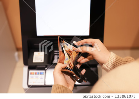 Hands holding a wallet with Euro banknotes and a credit card in front of an ATM screen, ready for a transaction. High quality photo Hands holding a wallet with Euro banknotes and a credit card in front of an ATM screen, ready for a transaction. High quality photo 133861075