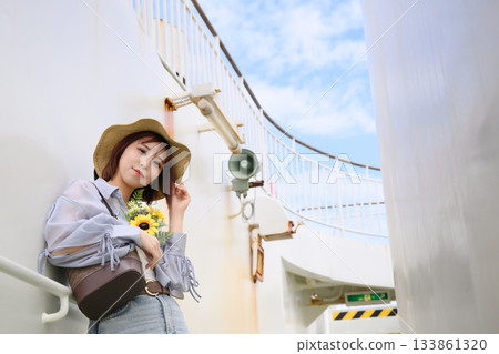 A young Japanese woman wearing a straw hat is posing on the deck of a summer ferry, holding a sunflower and with a bright blue sky in the background 133861320