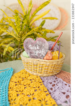 Basket of colorful yarn and crochet hooks and red wooden heart with Thank you lettering on background of surrounded colorful crocheted fabric and home flowers 133861947