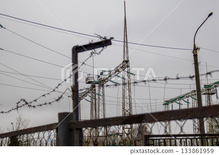 Barbed wire fences protect a tall electrical transmission tower in an urban area with gray skies overhead Barbed wire fences protect a tall electrical transmission tower in an urban area with gray skies overhead 133861959