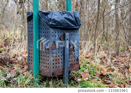 A metal trash bin, rusted and partially covered, stands in a forested location with dry leaves scattered around and trees in the background A metal trash bin, rusted and partially covered, stands in a forested location with dry leaves scattered around and trees in the background 133861961