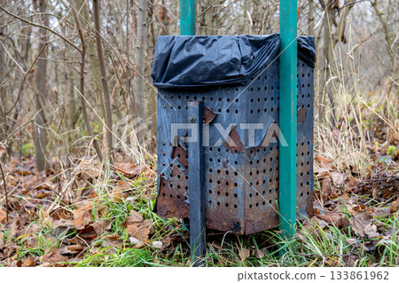 An old trash bin covered with a black bag stands in a forest clearing, surrounded by dried leaves and trees without foliage An old trash bin covered with a black bag stands in a forest clearing, surrounded by dried leaves and trees without foliage 133861962