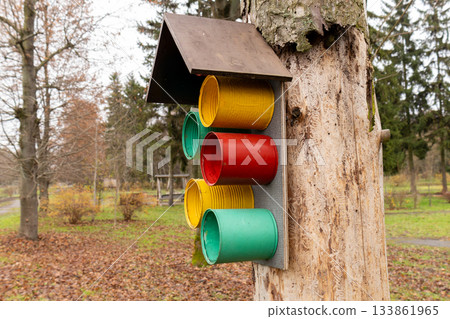 A bird feeder made of colorful canisters is fixed to a tree in a park. Leaves have fallen, and the ground is covered in autumn foliage A bird feeder made of colorful canisters is fixed to a tree in a park. Leaves have fallen, and the ground is covered in autumn foliage 133861965