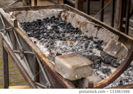 A rustic charcoal grill sits emptied, filled with ash and charcoal remnants after a lively gathering, capturing the essence of outdoor cooking 133861986