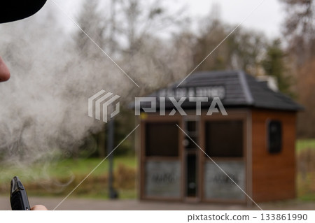 A person is vaping near a wooden booth in a park, releasing a cloud of vapor into the cool air as leaves change color nearby A person is vaping near a wooden booth in a park, releasing a cloud of vapor into the cool air as leaves change color nearby 133861990
