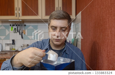 Man preparing a protein shake in a kitchen, measuring powder into a shaker for fitness, recovery, and muscle health. Man preparing a protein shake in a kitchen, measuring powder into a shaker for fitness, recovery, and muscle health. 133863324