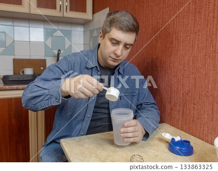 Man preparing a protein shake in a kitchen, measuring powder into a shaker for fitness, recovery, and muscle health. Man preparing a protein shake in a kitchen, measuring powder into a shaker for fitness, recovery, and muscle health. 133863325