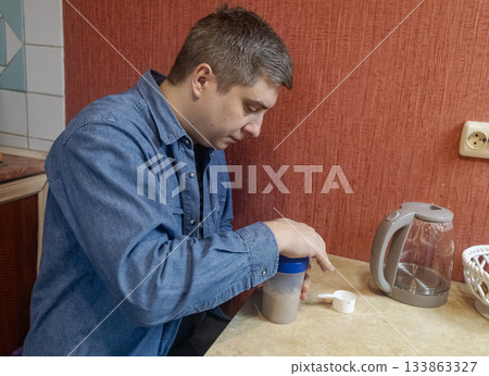 Man preparing a protein shake in a kitchen, measuring powder into a shaker for fitness, recovery, and muscle health. Man preparing a protein shake in a kitchen, measuring powder into a shaker for fitness, recovery, and muscle health. 133863327