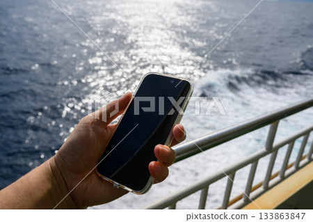 一名男子站在渡輪甲板上，手持智慧型手機，眺望著陽光灑在海面上的倒影。 133863847