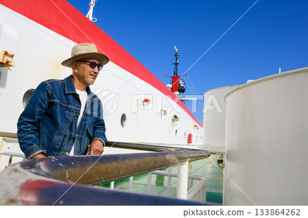 A Japanese man in his 50s wearing a straw hat and sunglasses looks out at the scenery on the deck of a ferry under a blue sky 133864262