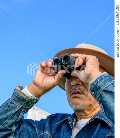 Senior man gazing into the distance through binoculars against the blue sky Senior man gazing into the distance through binoculars against the blue sky 133864289