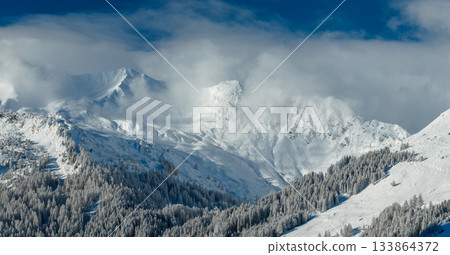 Panoramic photography of snowy alpine peaks in Austria with a glacier Hintertux rising through morning mist. Blue sky, sunlight, and conifer forests in the foreground create a peaceful winter 133864372