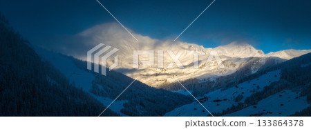 Panoramic photography of snowy alpine peaks in Austria with a glacier Hintertux rising through morning mist. Blue sky, sunlight, and conifer forests in the foreground create a peaceful winter 133864378