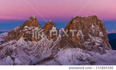 Panoramic view of Dolomite mountain peaks under a glowing full moon. Early winter landscape with snow-covered summits in soft dawn light. Panoramic view of Dolomite mountain peaks under a glowing full moon. Early winter landscape with snow-covered summits in soft dawn light. 133864380
