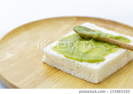 A close-up of a thick slice of white bread with Pandan Custard evenly applied on top, placed on a round wooden plate with a small 133865583