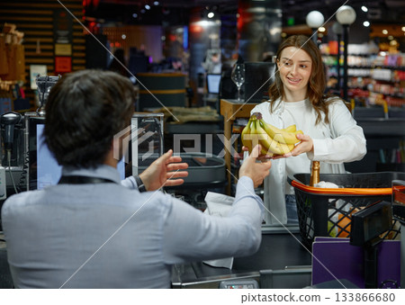 A woman is giving a banana to a man cashier at a grocery store 133866680