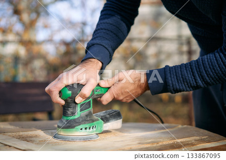 Close up of hands holding orbital sander while polishing wooden surface during renovation 133867095