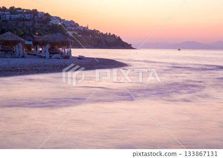 Thatched alcoves on the beach and white houses in distance at Greek islands  in pink sunset (maybe  Crete or  Santorini or  Spetsesd or Rhodes  or Mykonos 133867131