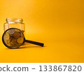 The image displays a clear glass jar filled with coins and a magnifying glass resting against it, set against a yellow background. 133867820